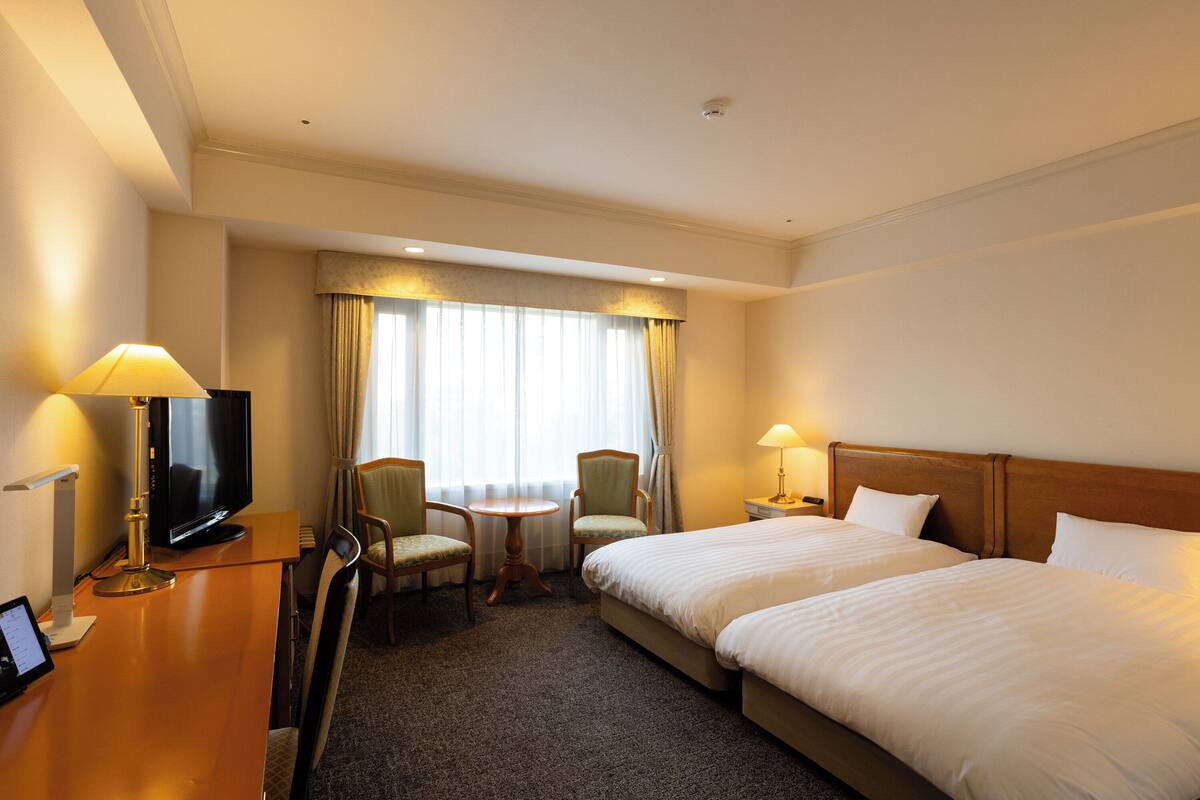 A hotel room with two beds, a TV, a desk with a lamp, and a small table with chairs.