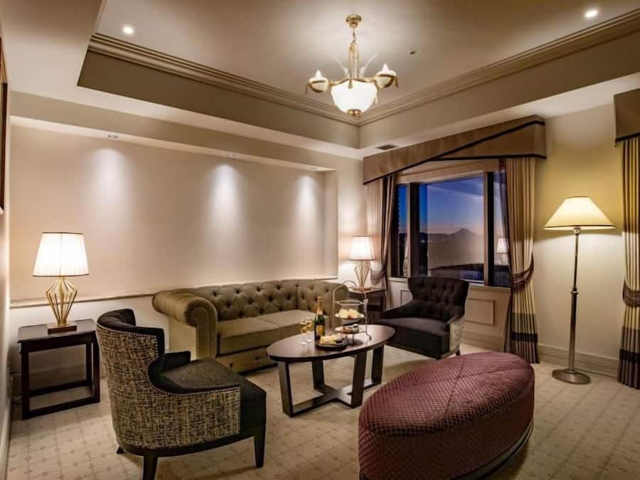 A hotel room with a sofa, armchairs, a coffee table, a floor lamp, and a view of the outdoors.