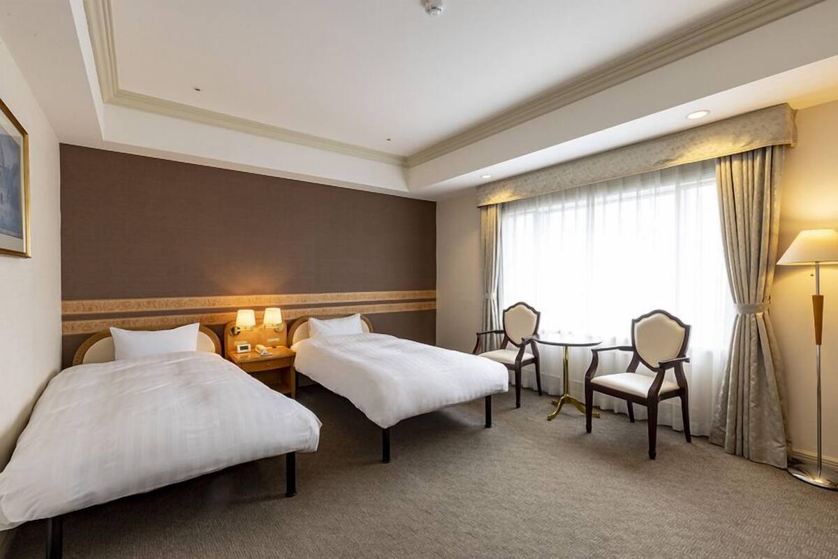 A hotel room with two beds, a small table, two chairs, a lamp, and a window with curtains.
