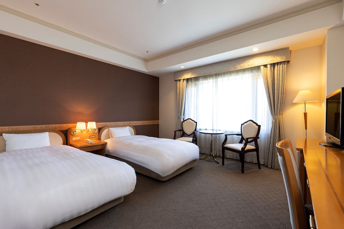 A hotel room with two beds, a desk, a chair, a small table, and a TV.