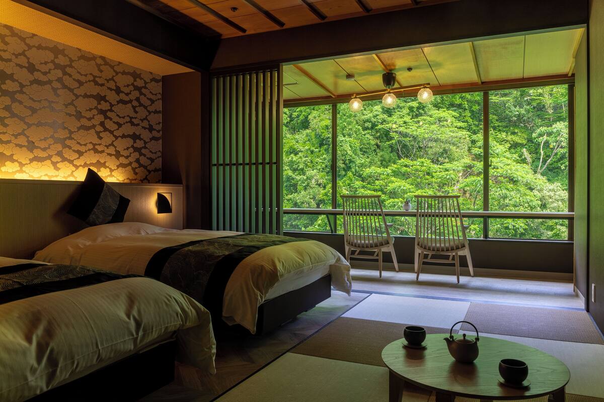 Une chambre avec deux lits, une vue sur une forêt grâce à de grandes fenêtres, un balcon avec une chaise et une petite table avec une théière et une tasse.