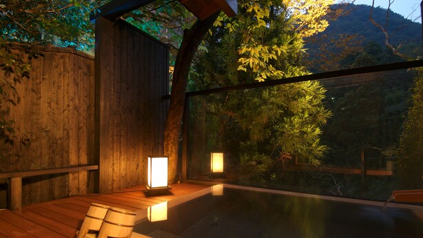 Une terrasse en bois avec un jacuzzi et deux lanternes, entourée d’arbres et offrant une vue sur la montagne.