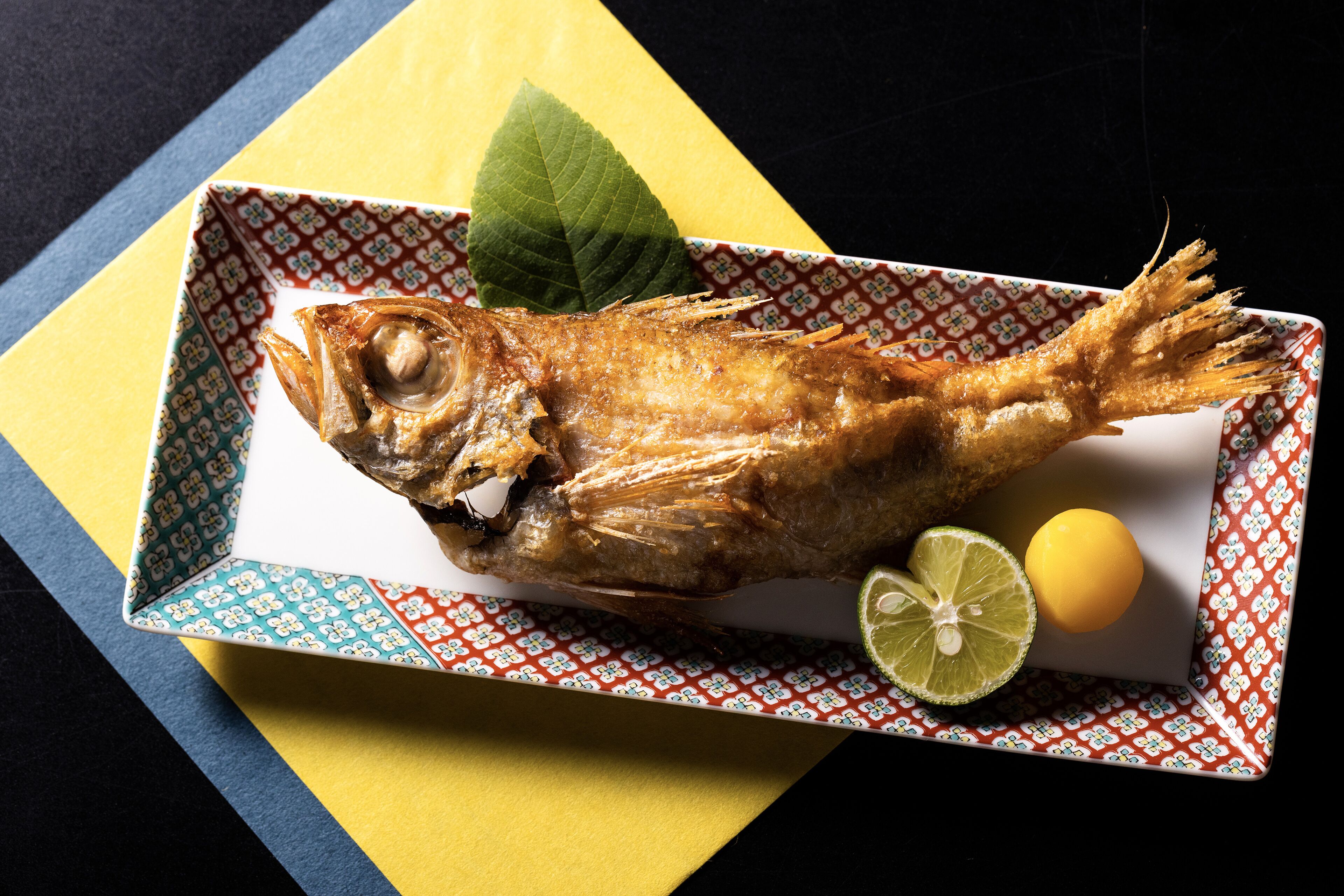 Un pescado entero frito sobre un plato estampado, con un gajo de lima y media limón.