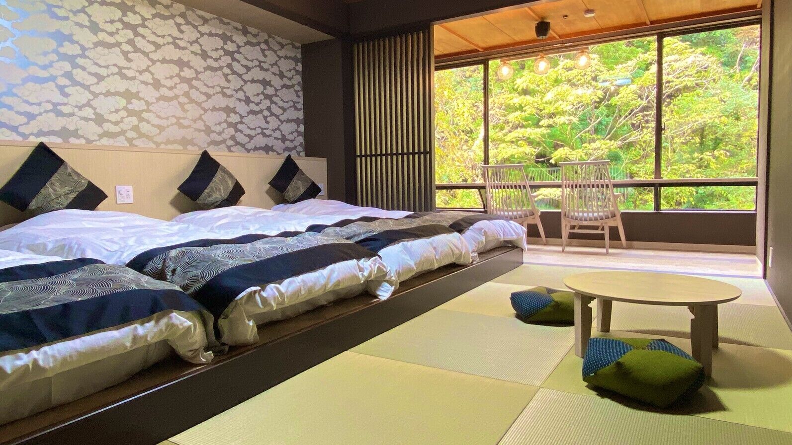 A room with a bed, a small table, and a view of greenery through large windows.