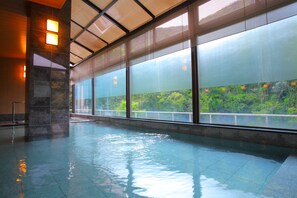 An indoor swimming pool with clear glass walls, a tiled floor, and a view of a green landscape. - Yamanaka Onsen Ohanami Kyubei (Kaga)