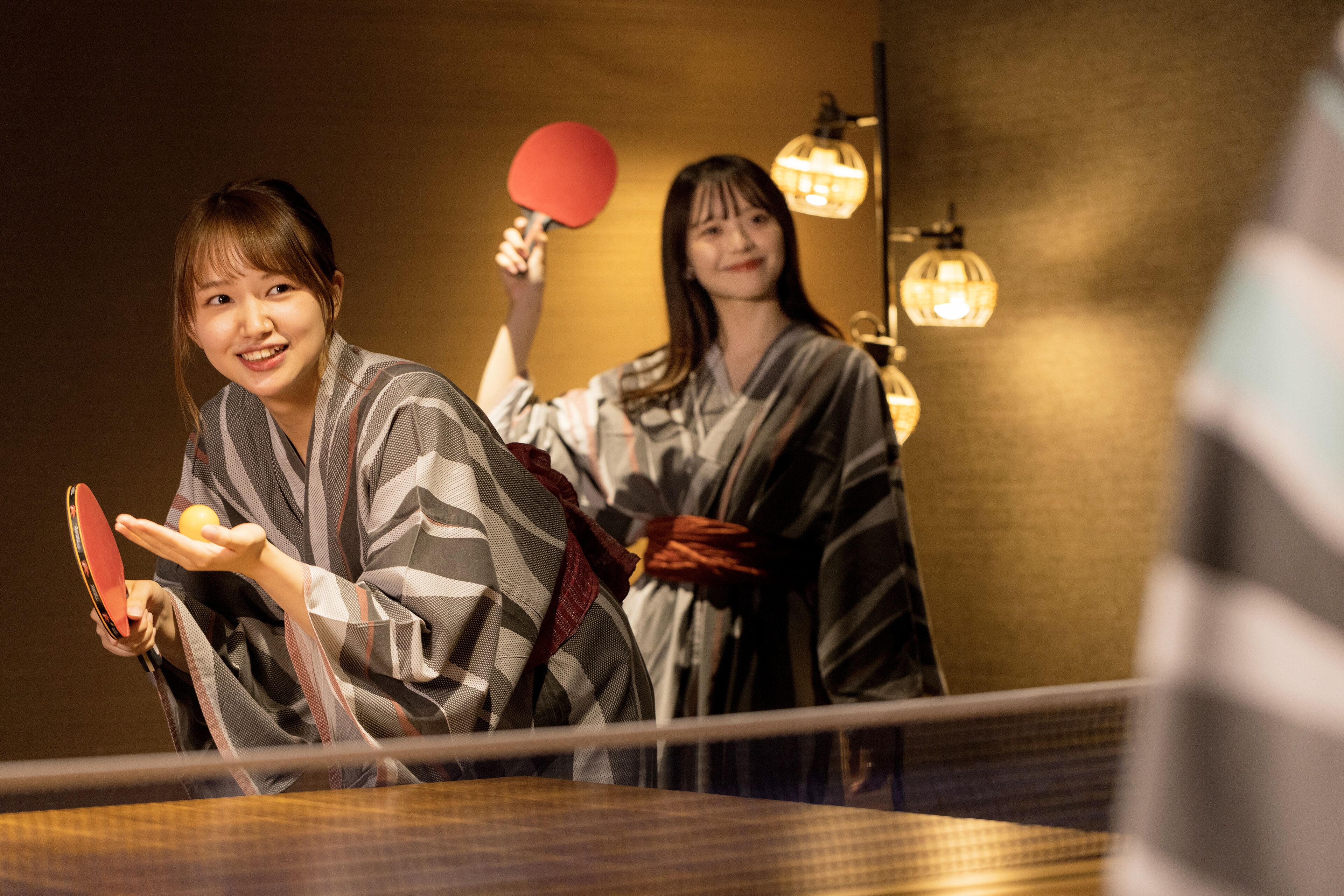 Two people in traditional attire playing table tennis in a room with a wooden table and hanging lamps.