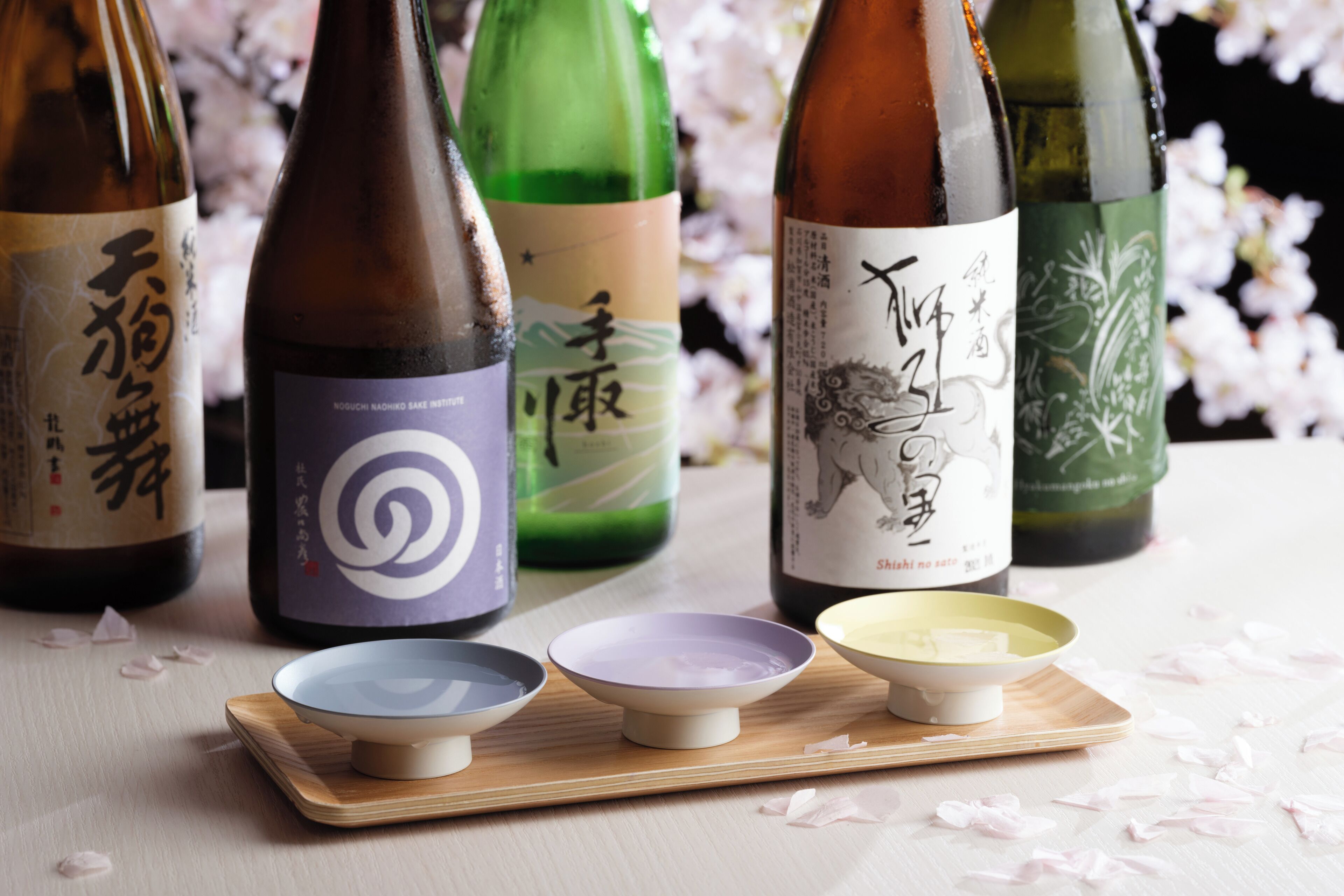 A selection of sake bottles with labels, arranged on a wooden tray with three small bowls in front.