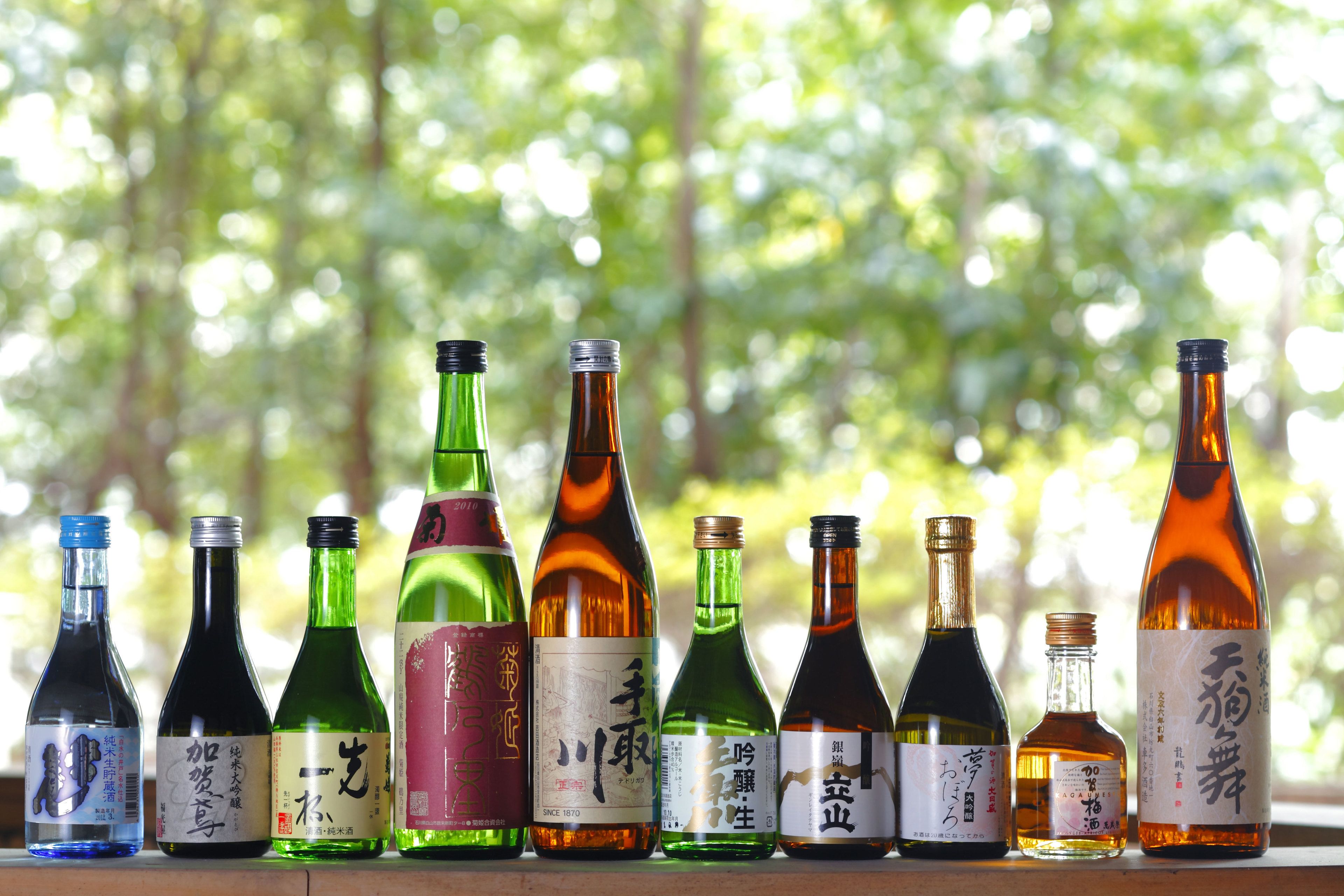 A row of variously shaped and colored bottles with labels, set against a blurred green background.