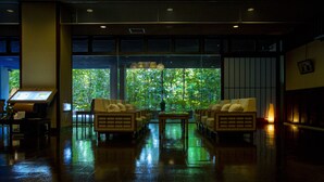 A spacious lobby with a wet floor, seating area with cushioned chairs, a reception desk, and large windows offering a view of greenery outside. - Fujiya Ryokan (Kaga)