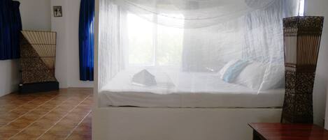 A bed with a canopy, a wooden table, and a chair in a room with tiled flooring and curtains.