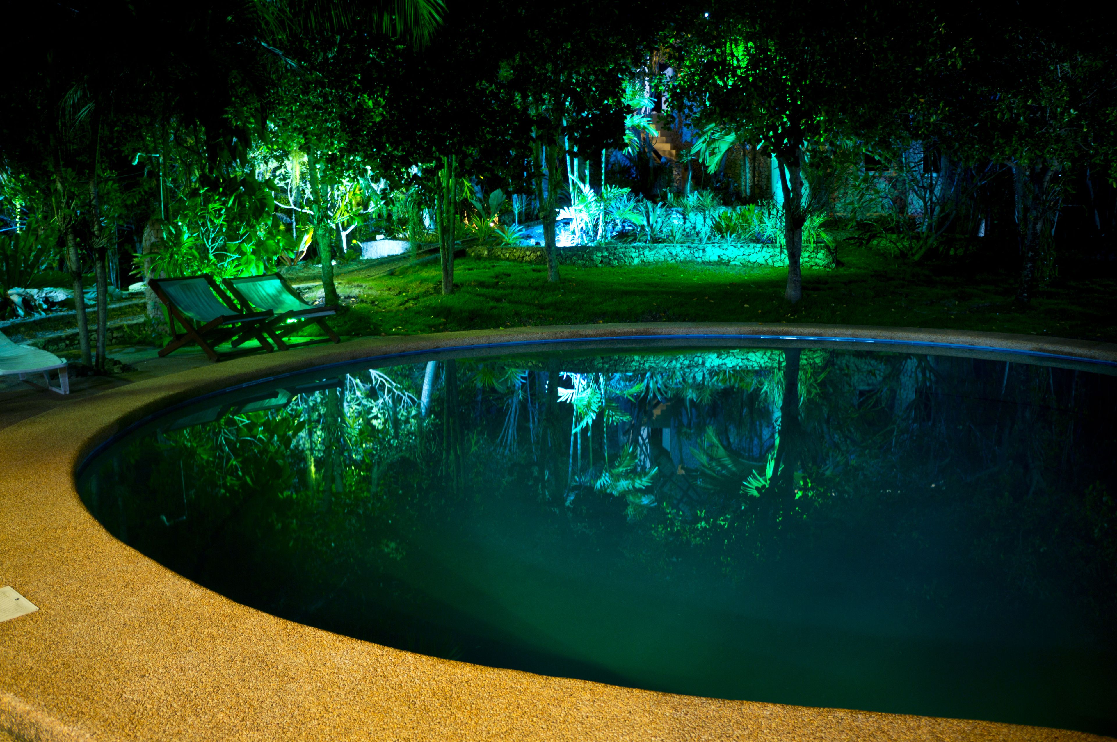A nighttime view of a swimming pool with green lighting, surrounded by trees and a grassy area with lounge chairs.