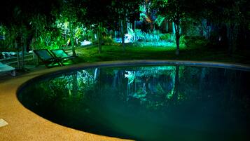A nighttime view of a swimming pool with green lighting, surrounded by trees and a grassy area with lounge chairs.
