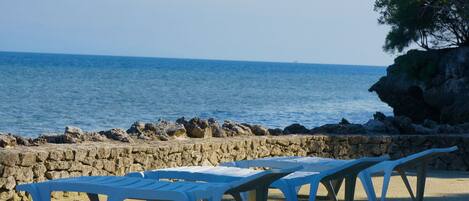 A row of white plastic beach chairs on a sandy beach with a stone wall and rocky outcrop in the background.