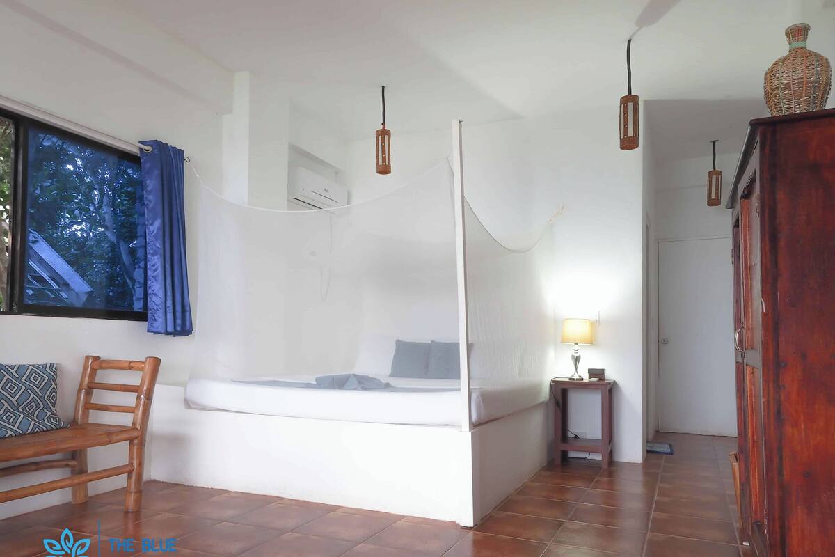 A bedroom with a bed, a bedside table, a chair, a window with curtains, and a view of trees outside.