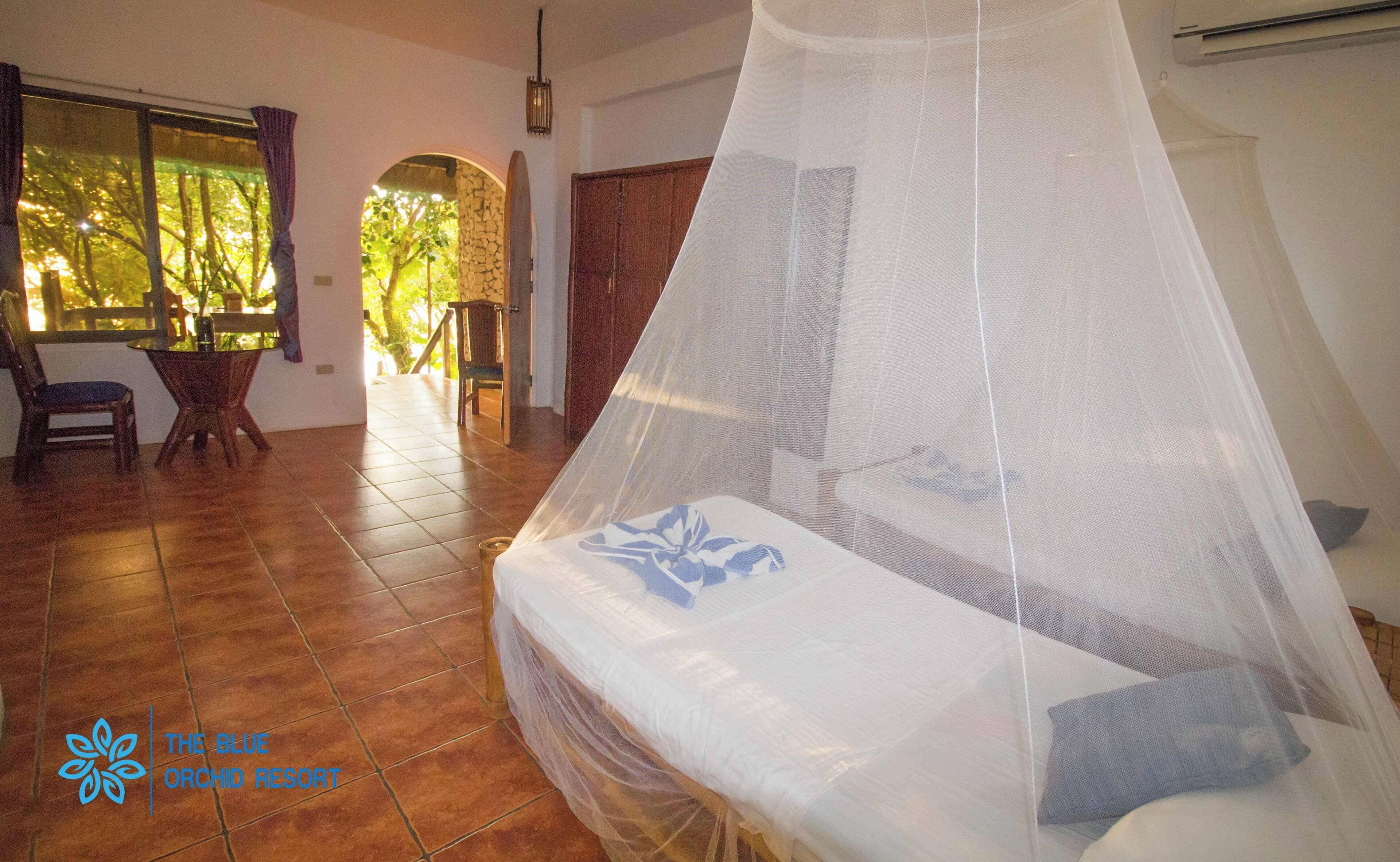 A bedroom with a bed under a mosquito net, a window with curtains, a chair, and a table.