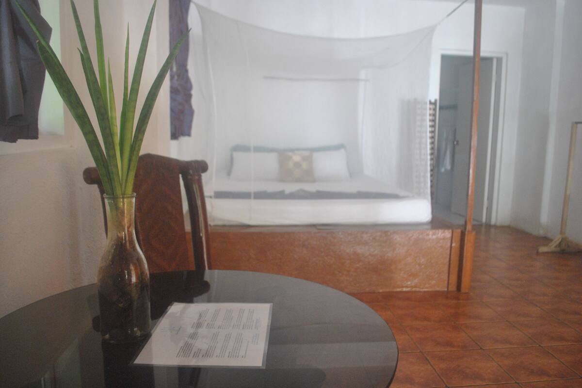 A room with a bed, a glass table with a vase, and a chair.