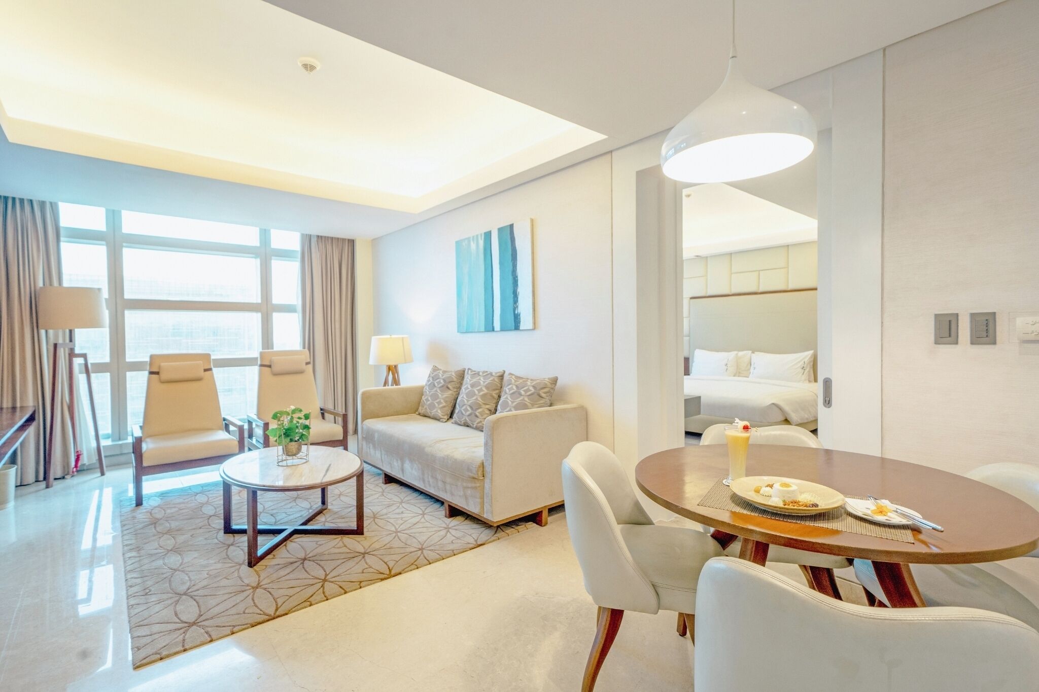 A modern hotel room with a sofa, two armchairs, a dining table, and a large window.