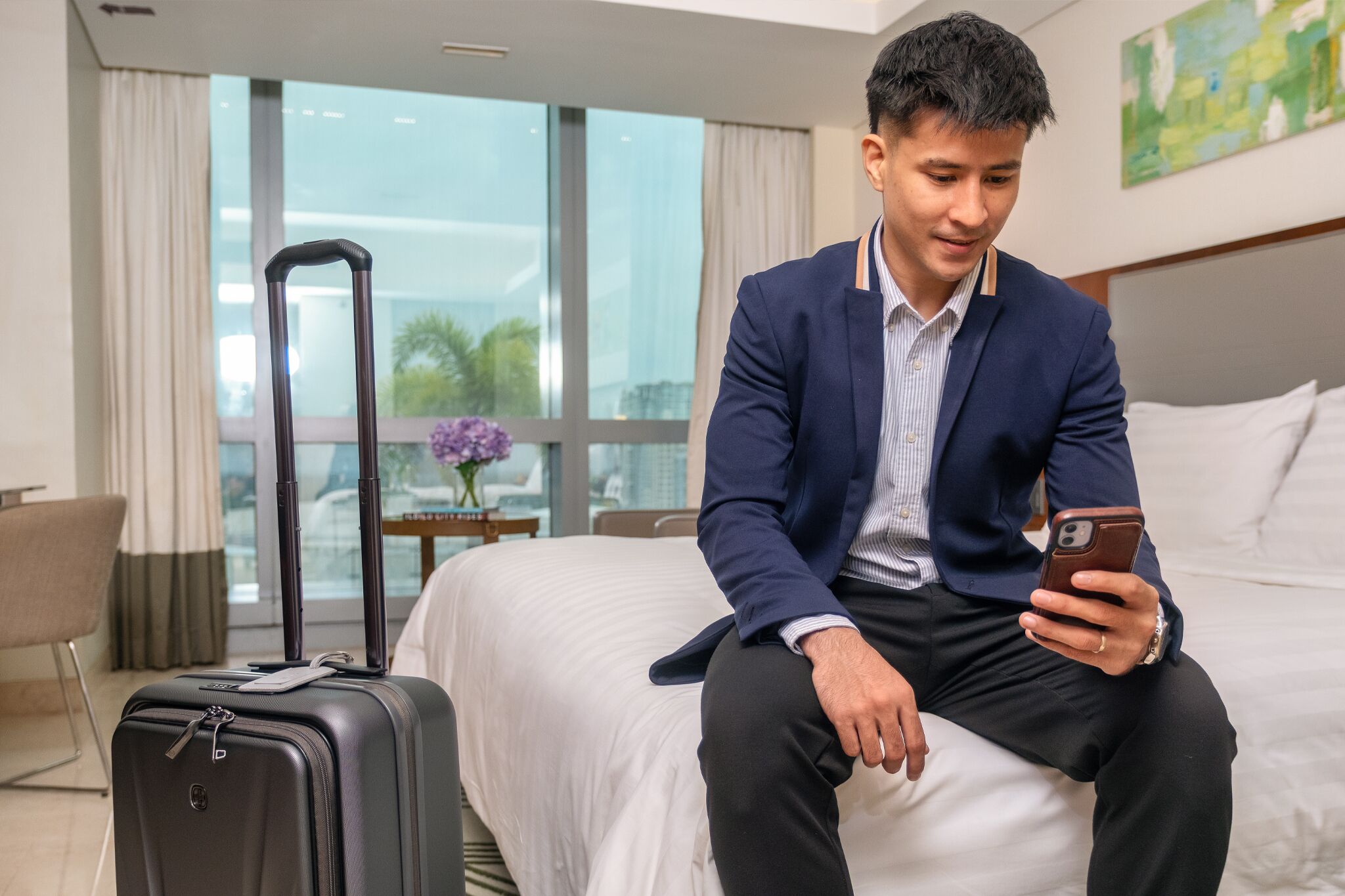 Une chambre d’hôtel avec un lit, une valise et une personne assise sur le lit, tenant un téléphone.