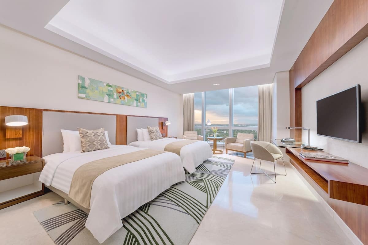 A modern hotel room with two beds, a desk, a TV, and a large window offering a city view.