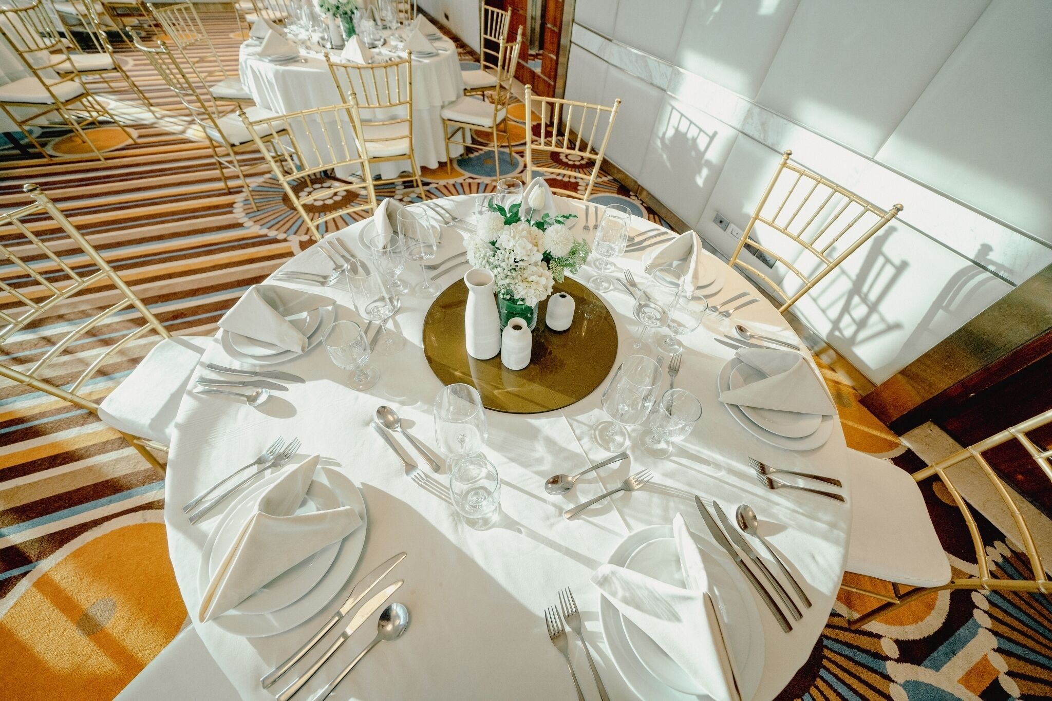 Une salle de banquet avec des tables rondes dressées pour un événement formel, agrémentée de touches dorées et recouverte de nappes blanches.