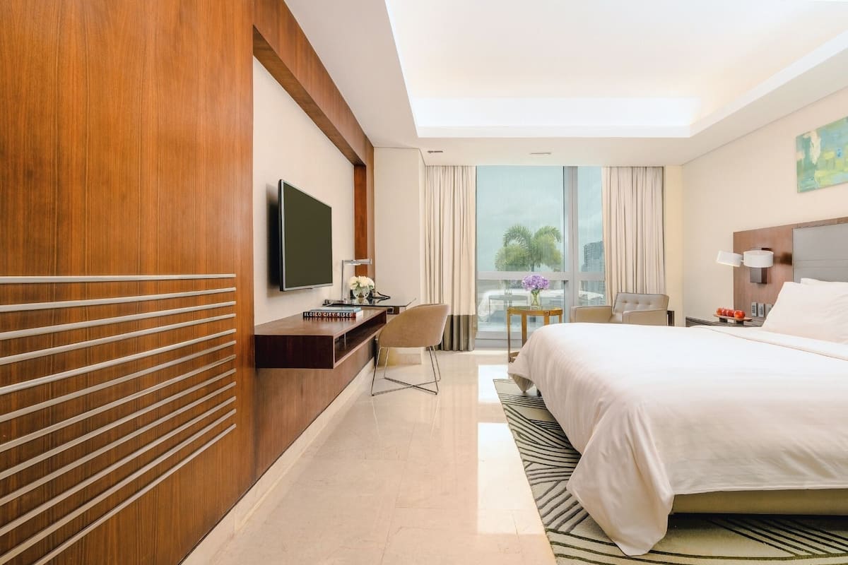 A hotel room with a large bed, a desk with a flat-screen TV, a chair, a small table with a vase of flowers, and a view of palm trees through the window.
