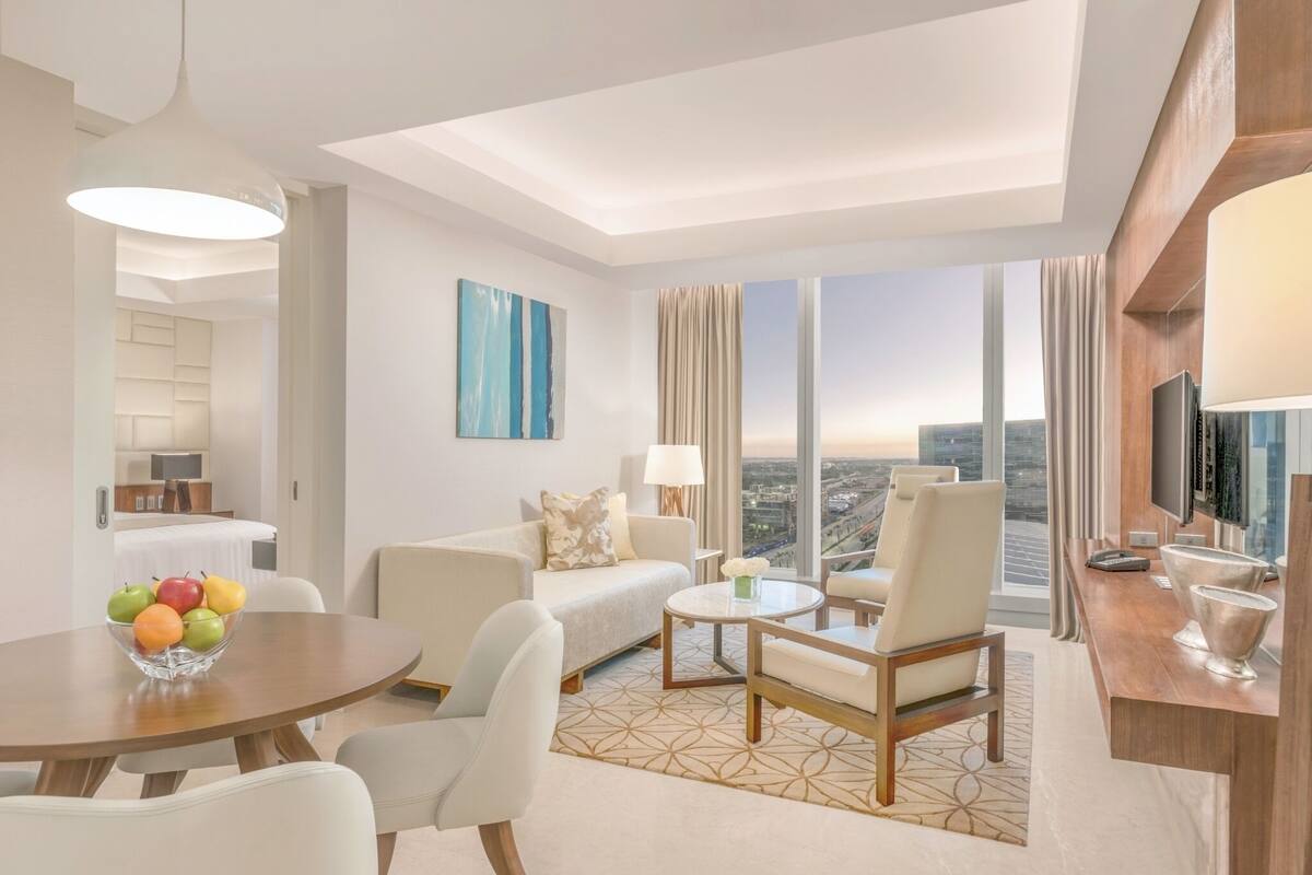A modern hotel room with a dining area, a sofa set, a TV, and a city view.
