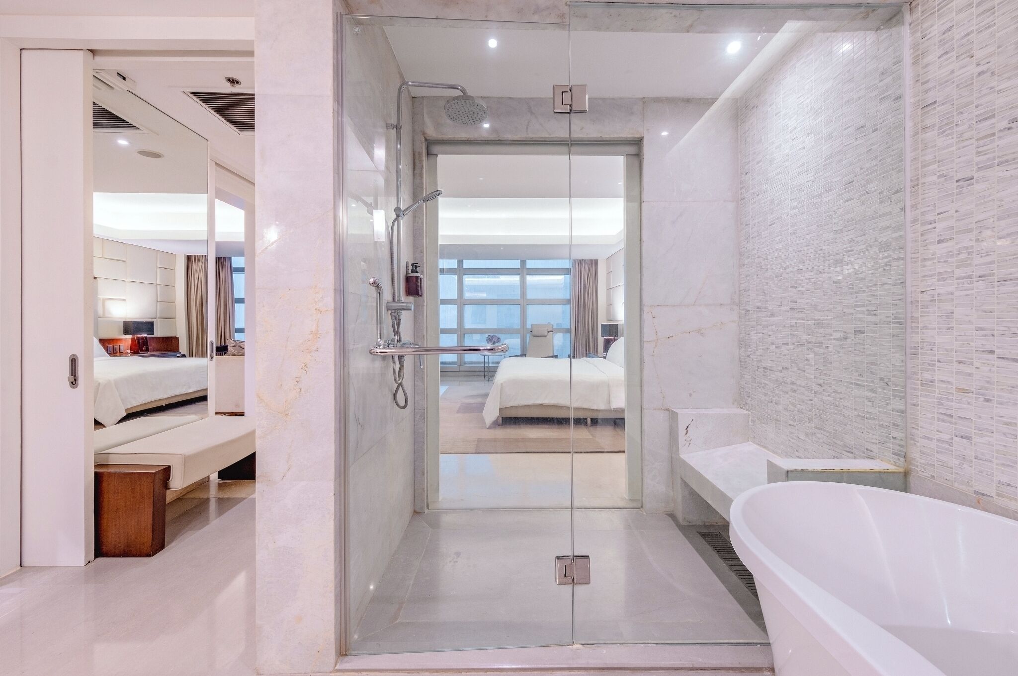 Un baño moderno con ducha de vidrio, una bañera y se ve un dormitorio con cama y mesita de noche.