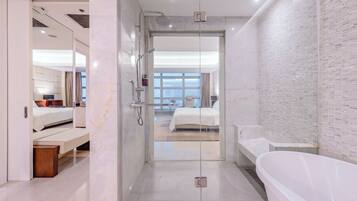 Un baño moderno con ducha de vidrio, una bañera y se ve un dormitorio con cama y mesita de noche.