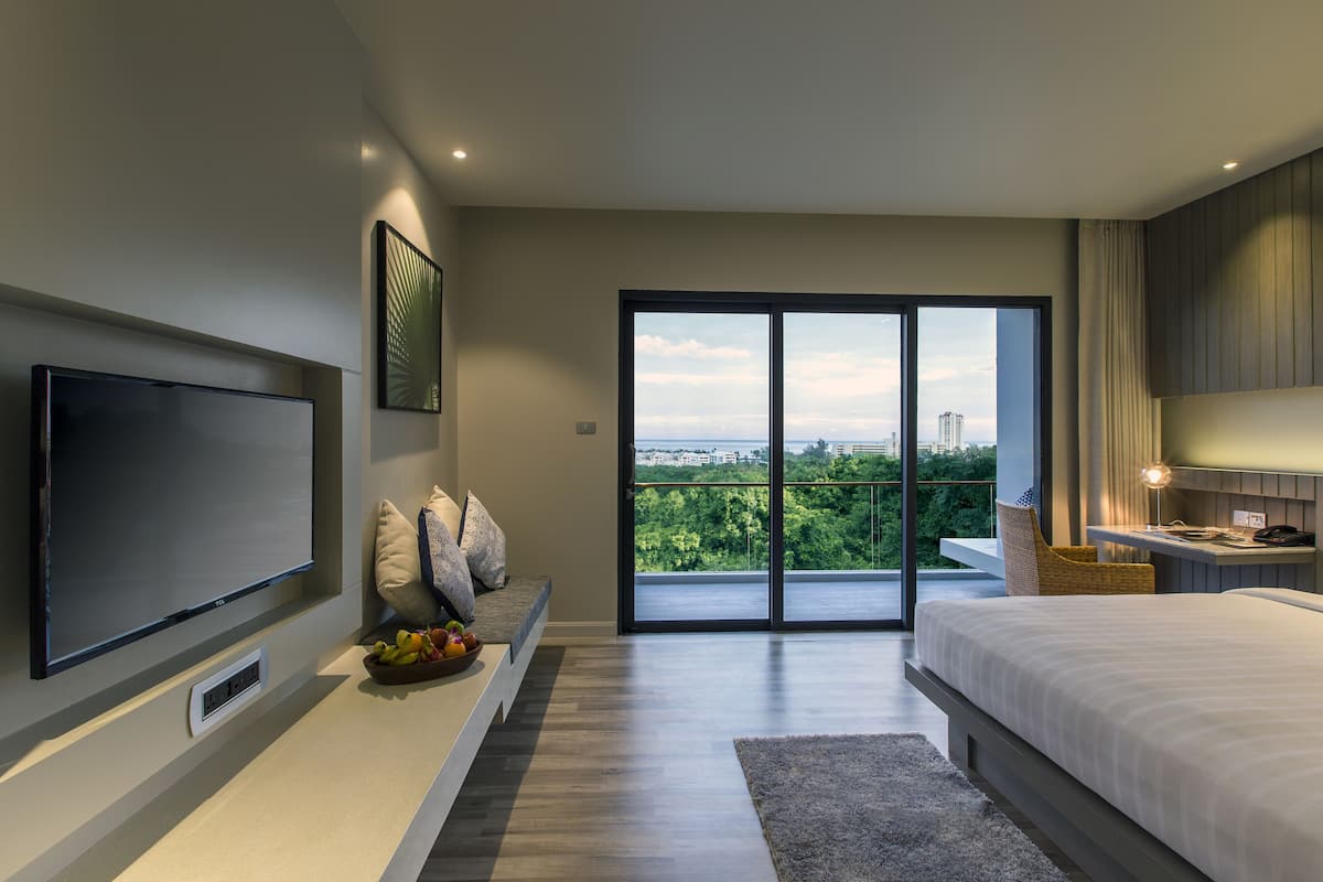 Une chambre moderne dotée d’un grand lit, d’un téléviseur à écran plat, d’un coin repas avec un panier de fruits et d’un balcon offrant une vue sur la ville.