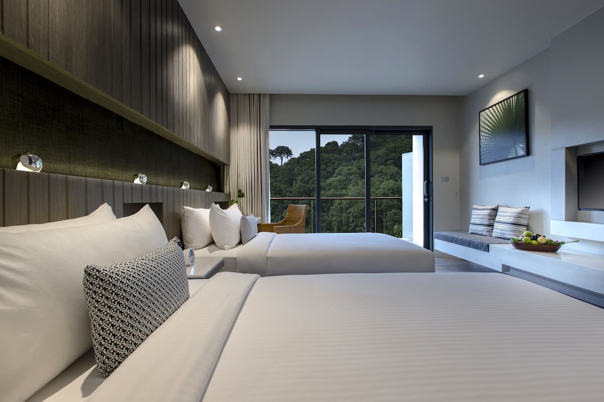 Une chambre d’hôtel moderne avec deux lits, une grande fenêtre donnant sur la verdure, un téléviseur fixé au mur et un coin salon agrémenté d’un panier de fruits.