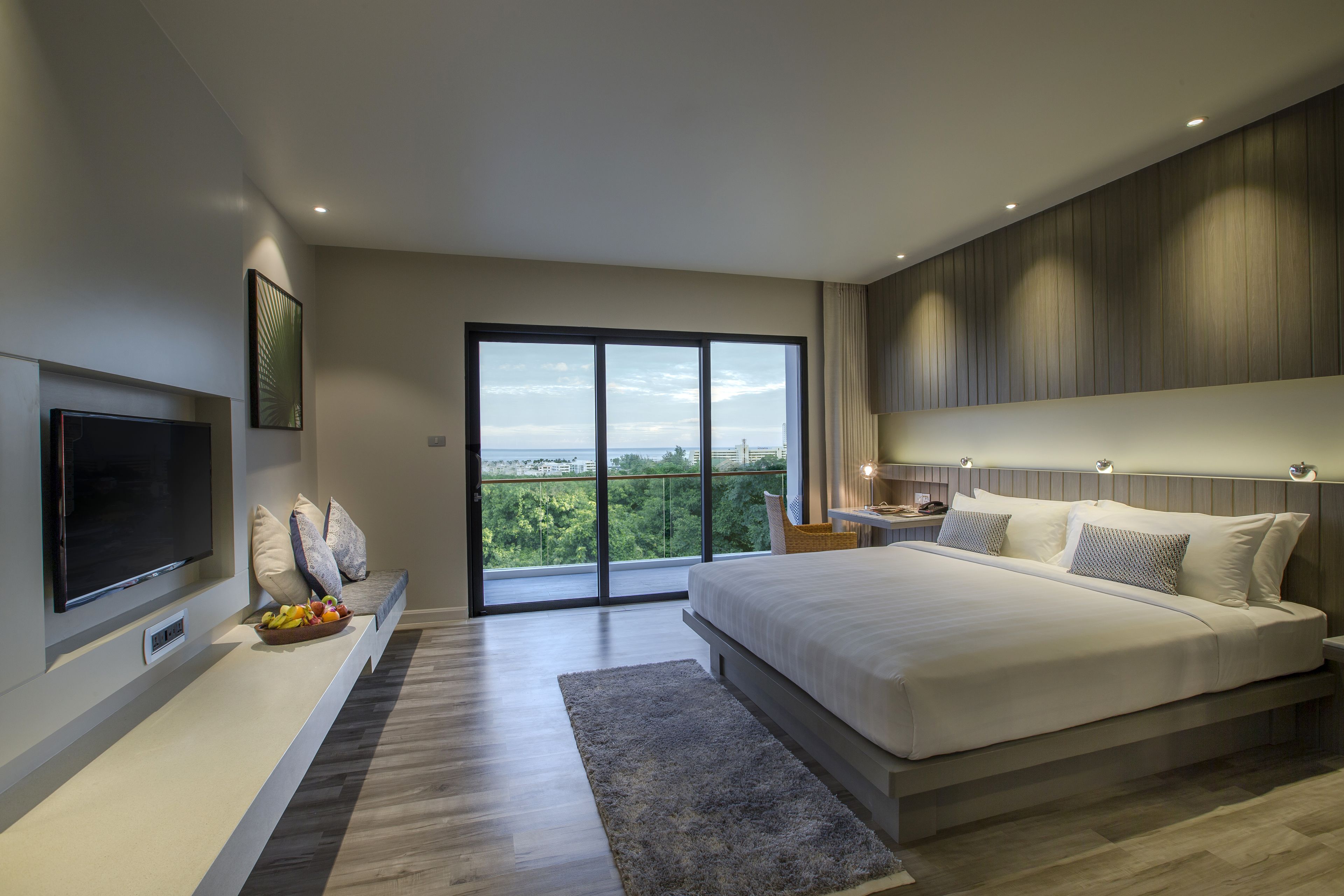 A modern bedroom with a large bed, a flat-screen TV, and a balcony with a city view.