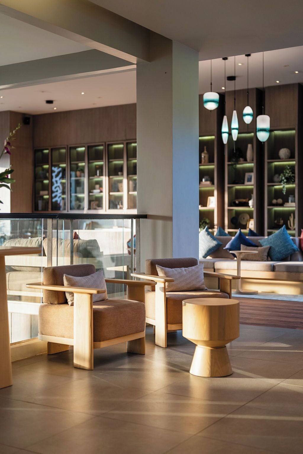 A modern lounge area with wooden furniture, a glass railing, and a view of a restaurant with illuminated windows.