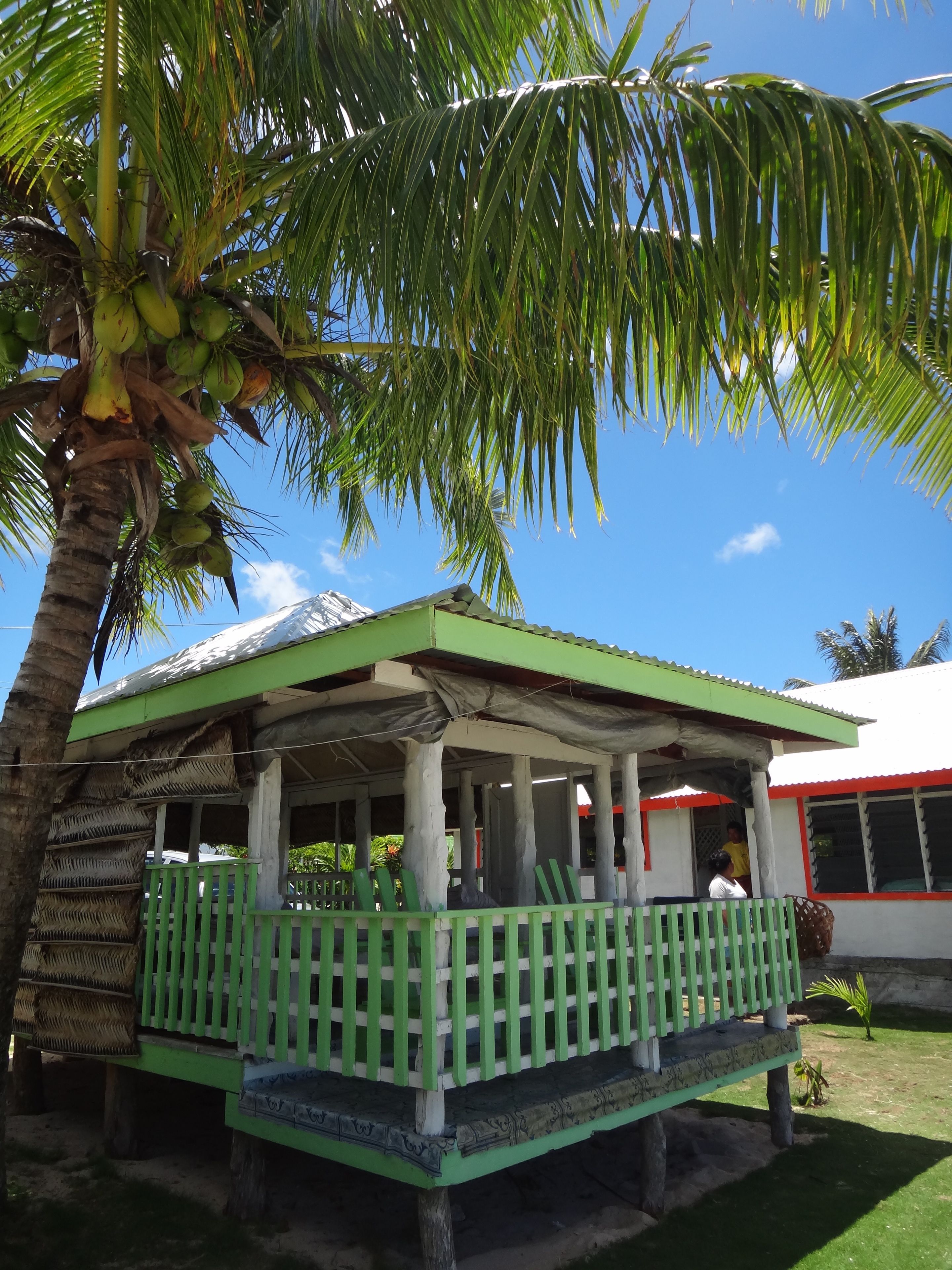 A small, single-story structure with a green roof and white walls, surrounded by palm trees and a clear blue sky.