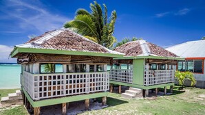 Dos cabañas de techo de paja con barandillas blancas, ubicadas en una playa de arena con cielo azul claro y palmeras al fondo.