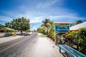 A beachside road with a sign welcoming to Tailua Beach Fales, a bench, and a small structure with a green roof. - Tailua Beach Fale (Manase)