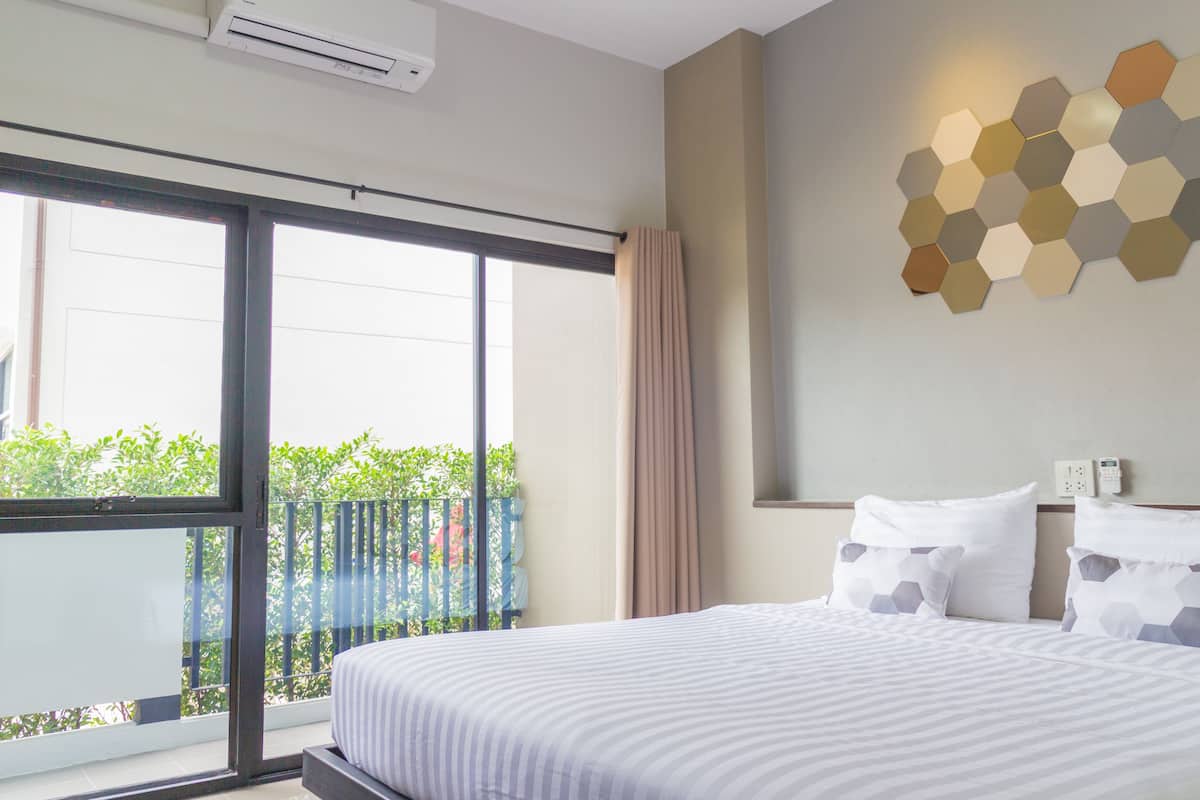A hotel room with a large bed, a view of greenery through the window, and a wall-mounted hexagonal light fixture.