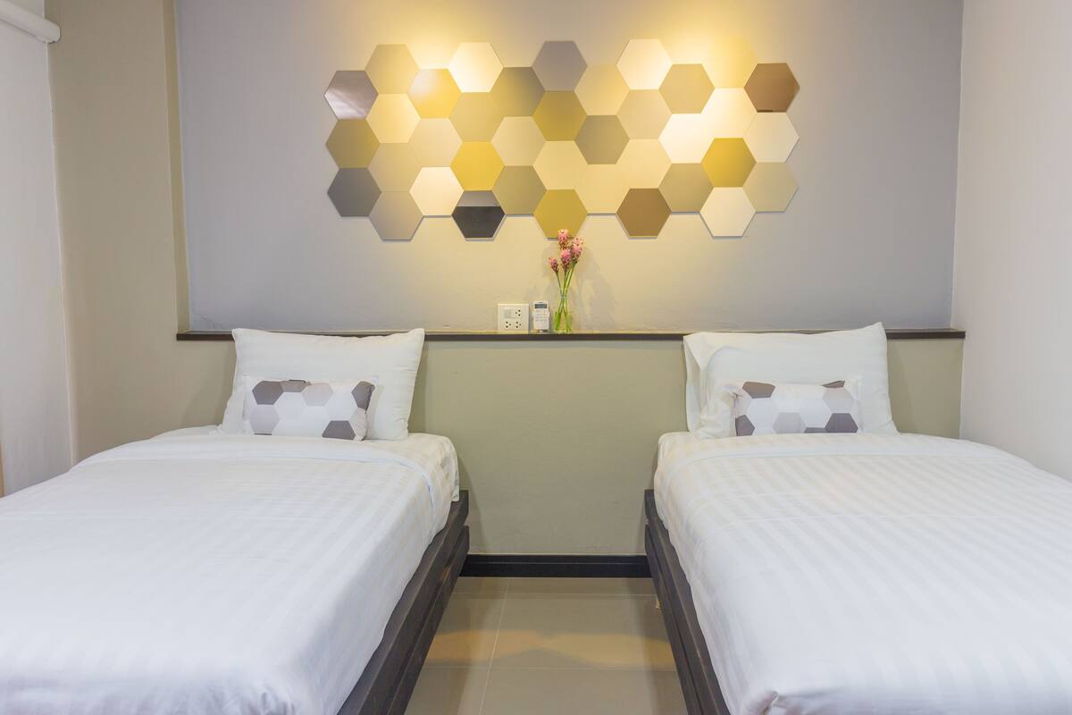 A hotel room with two beds, a wall-mounted hexagonal light fixture, a vase with flowers, and a small table with a telephone and power outlet.