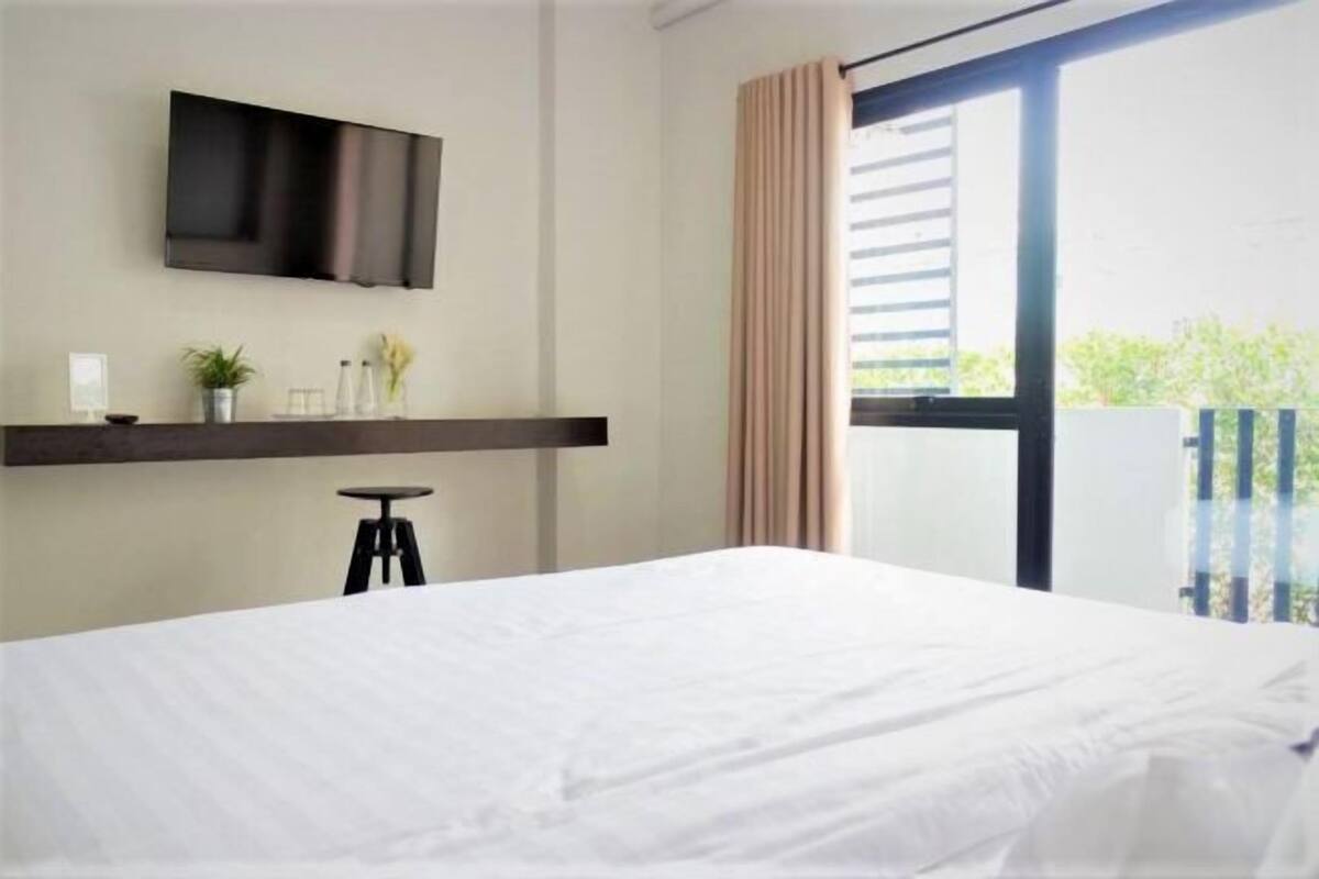 A hotel room with a bed, a mounted TV, a small table with a chair, a window with curtains, and a balcony view.