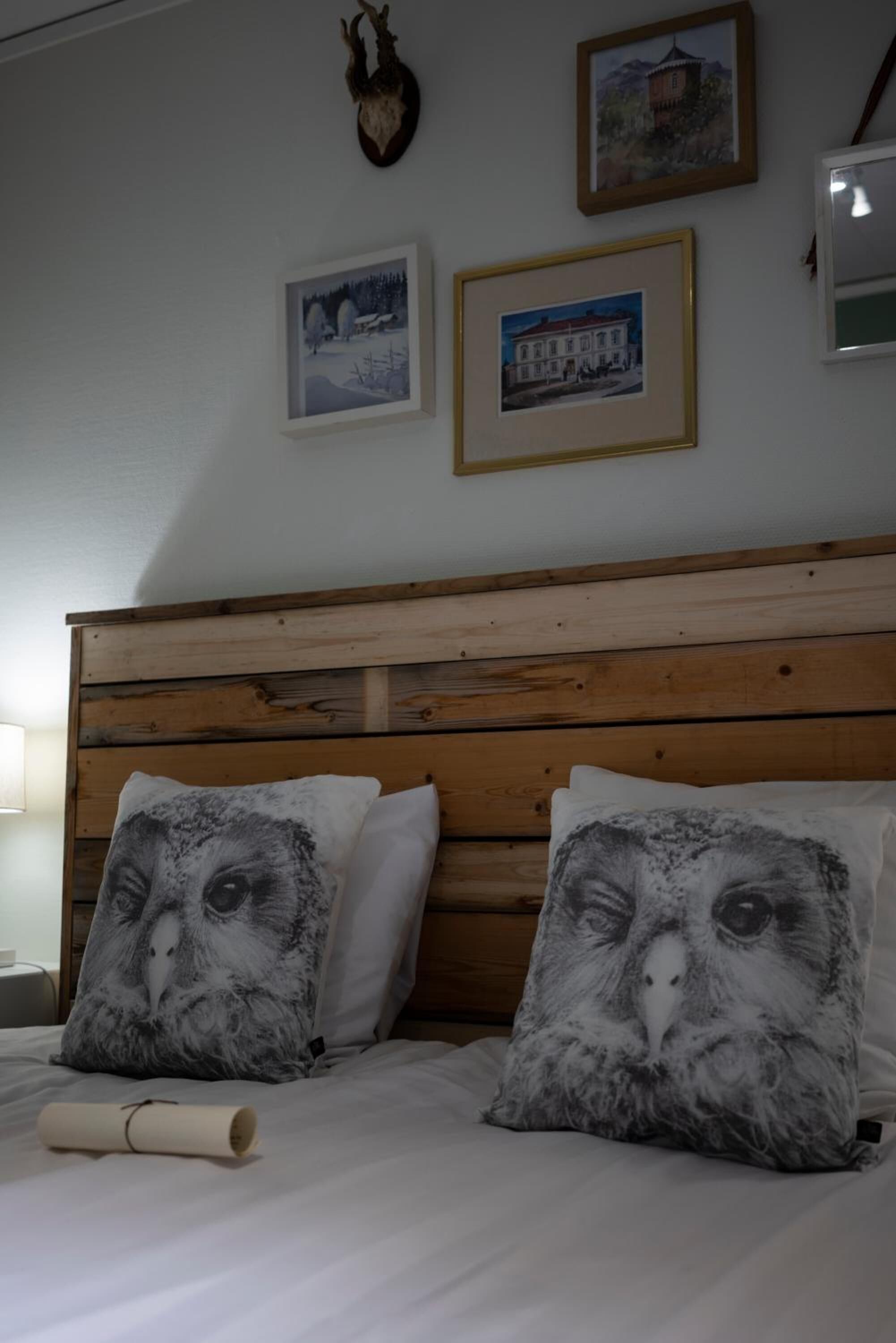 Un dormitorio con cabecera de madera, dos almohadas con estampados de lechuzas y cuadros en la pared.