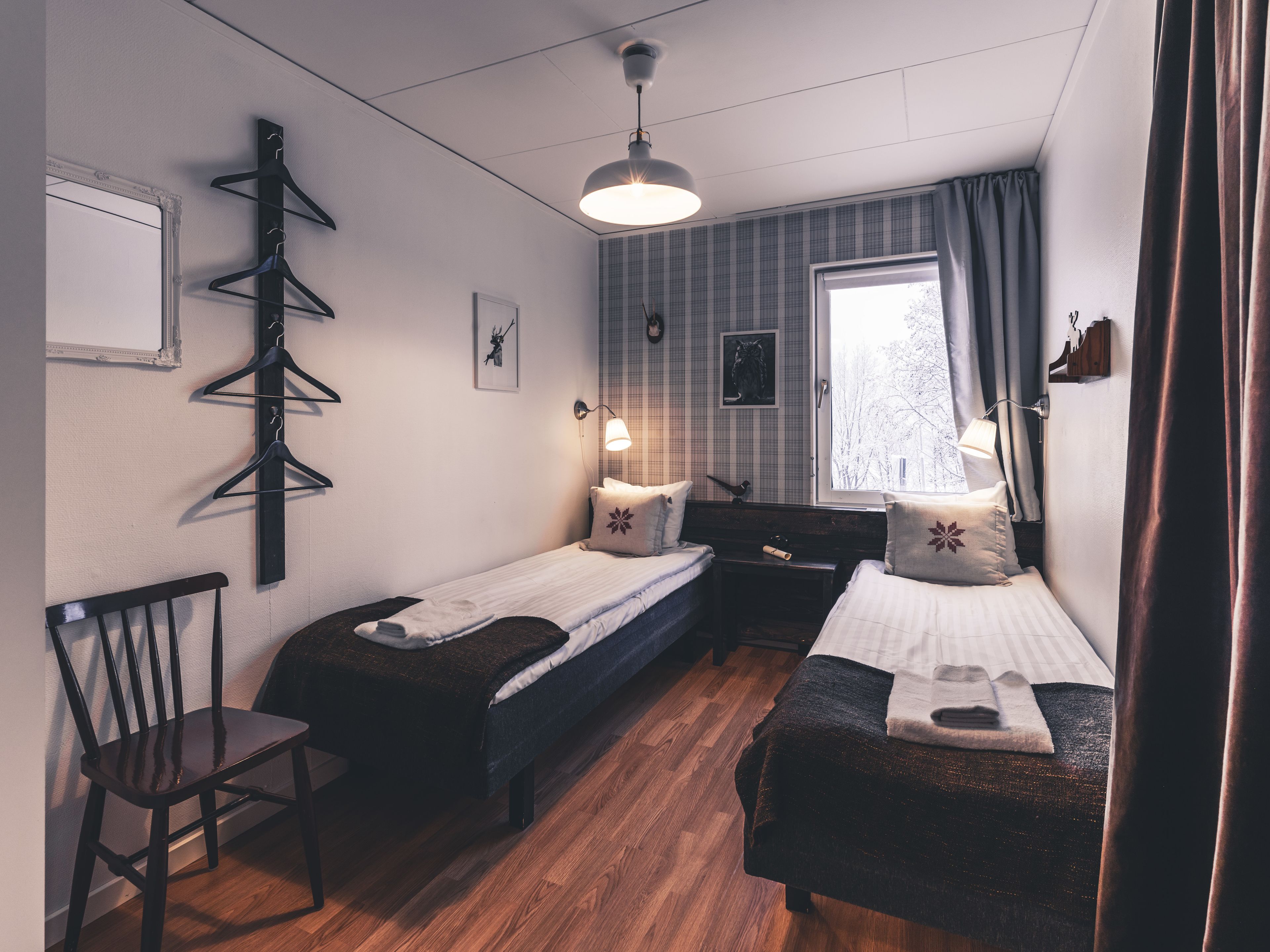 Quarto compacto com duas camas de solteiro, uma cadeira de madeira, escrivaninha e janela com cortinas.