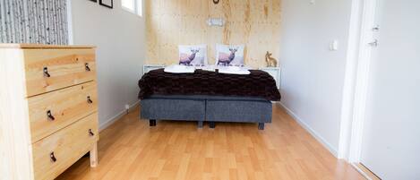 A bedroom with a wooden floor, a bed with a dark blanket, a wooden dresser, and a wall-mounted coat rack.