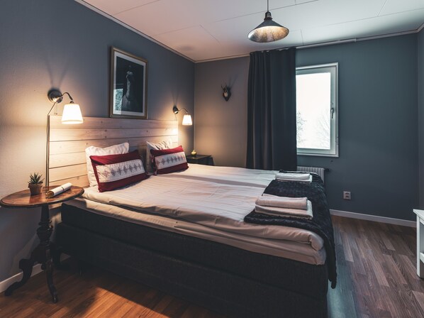 A hotel room with a double bed, two bedside lamps, a small table with a plant, a framed picture on the wall, and a window with curtains.