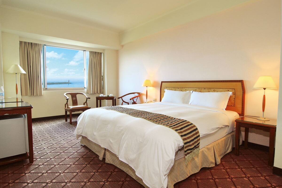 Una habitación de hotel con una cama grande, dos mesitas de noche, un escritorio, una silla y una ventana con cortinas.