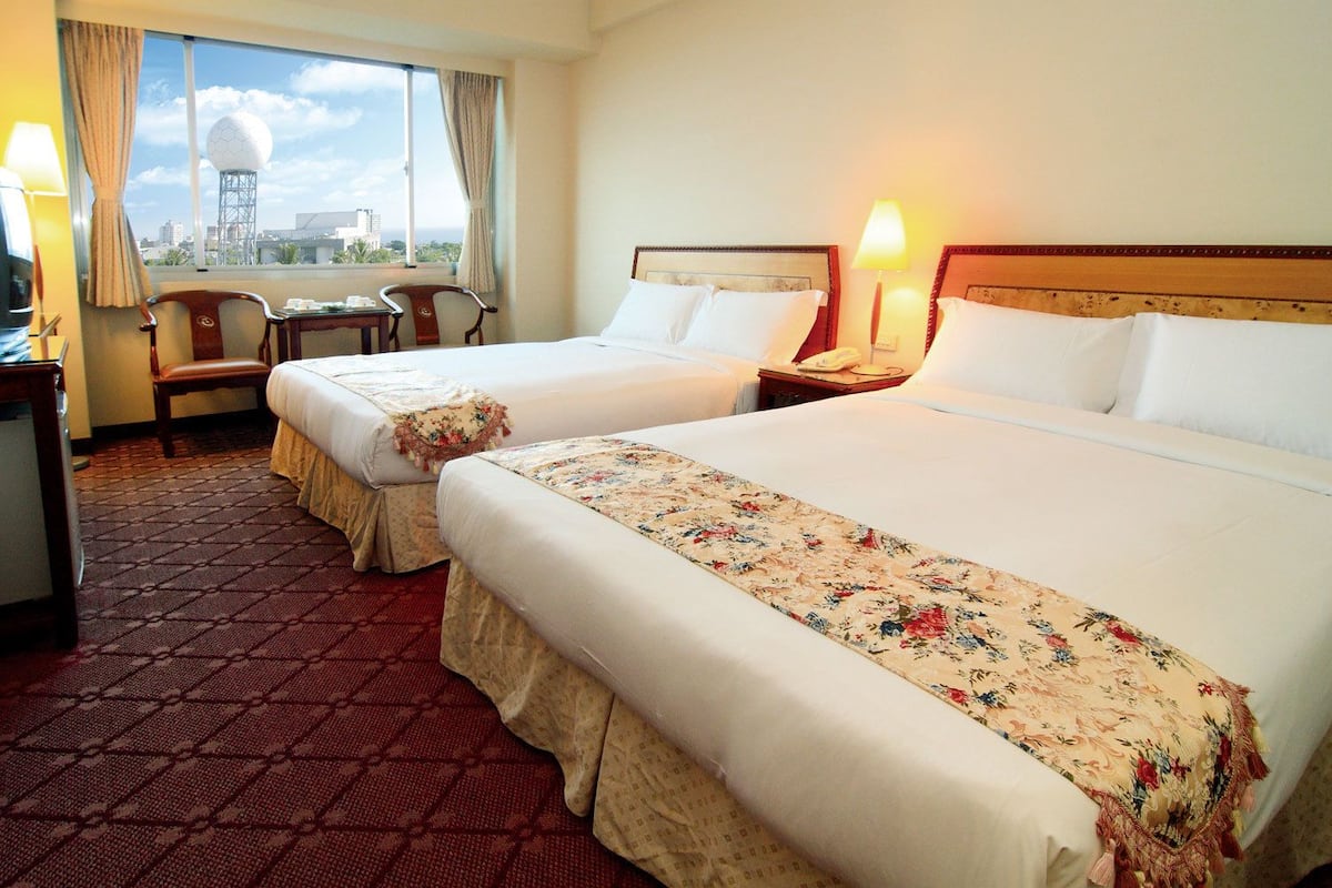 Habitación de hotel con dos camas, un escritorio, una silla y vistas a un paisaje urbano.