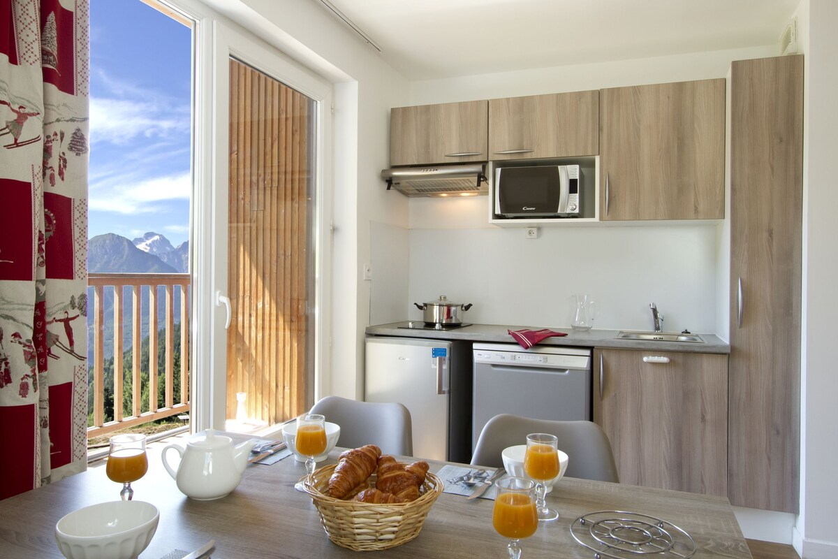 A compact kitchen with a microwave, a small refrigerator, and a sink. There is a dining table with a basket of croissants, a teapot, and glasses of orange juice.