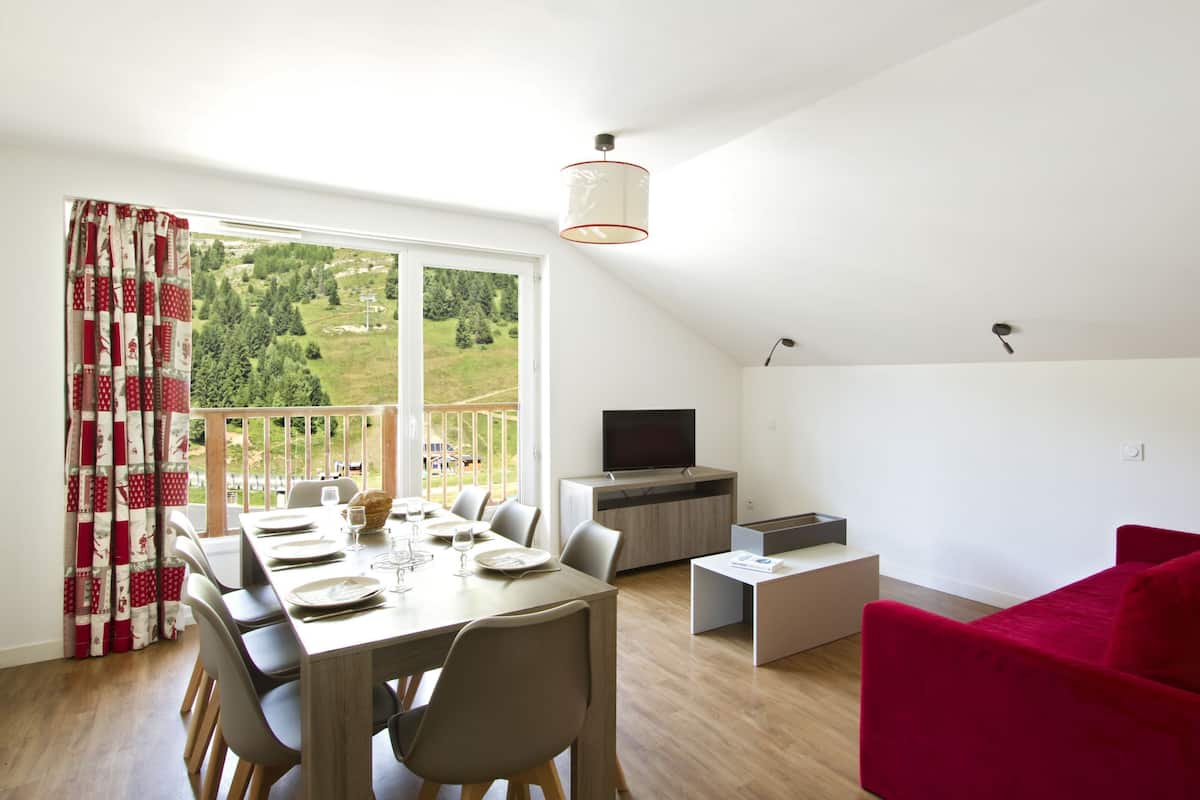 A modern living room with a dining area, a red sofa, a TV, and a balcony with a scenic view.