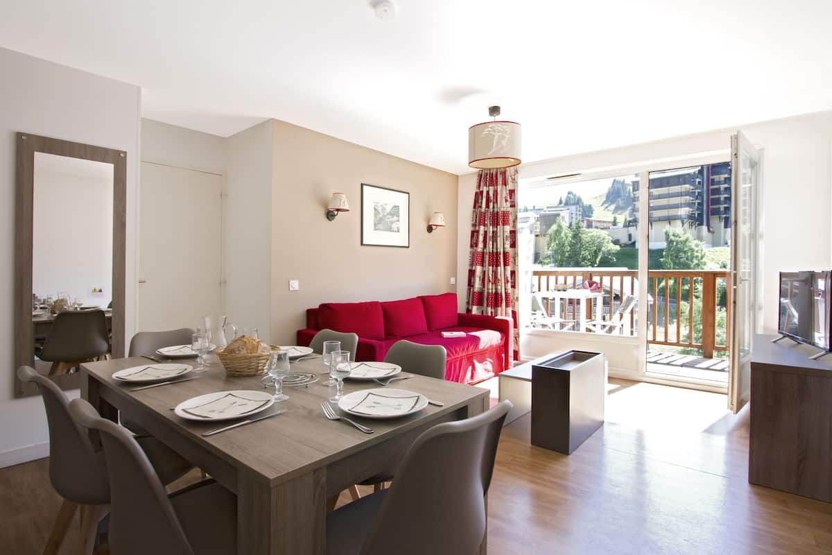A dining area with a wooden table set for four, a red sofa, and a balcony with a view of buildings and greenery.