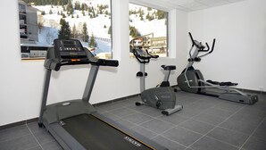 Et fitnesscenter med løbebånd, crosstrainer og motionscykel, med store vinduer og udsigt til et snækkedækket bjerg.