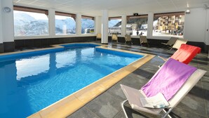 En indendørs swimmingpool med krystalklare udsigter til bjerge gennem store vinduer, liggestole med håndklæder og et flisegulv.