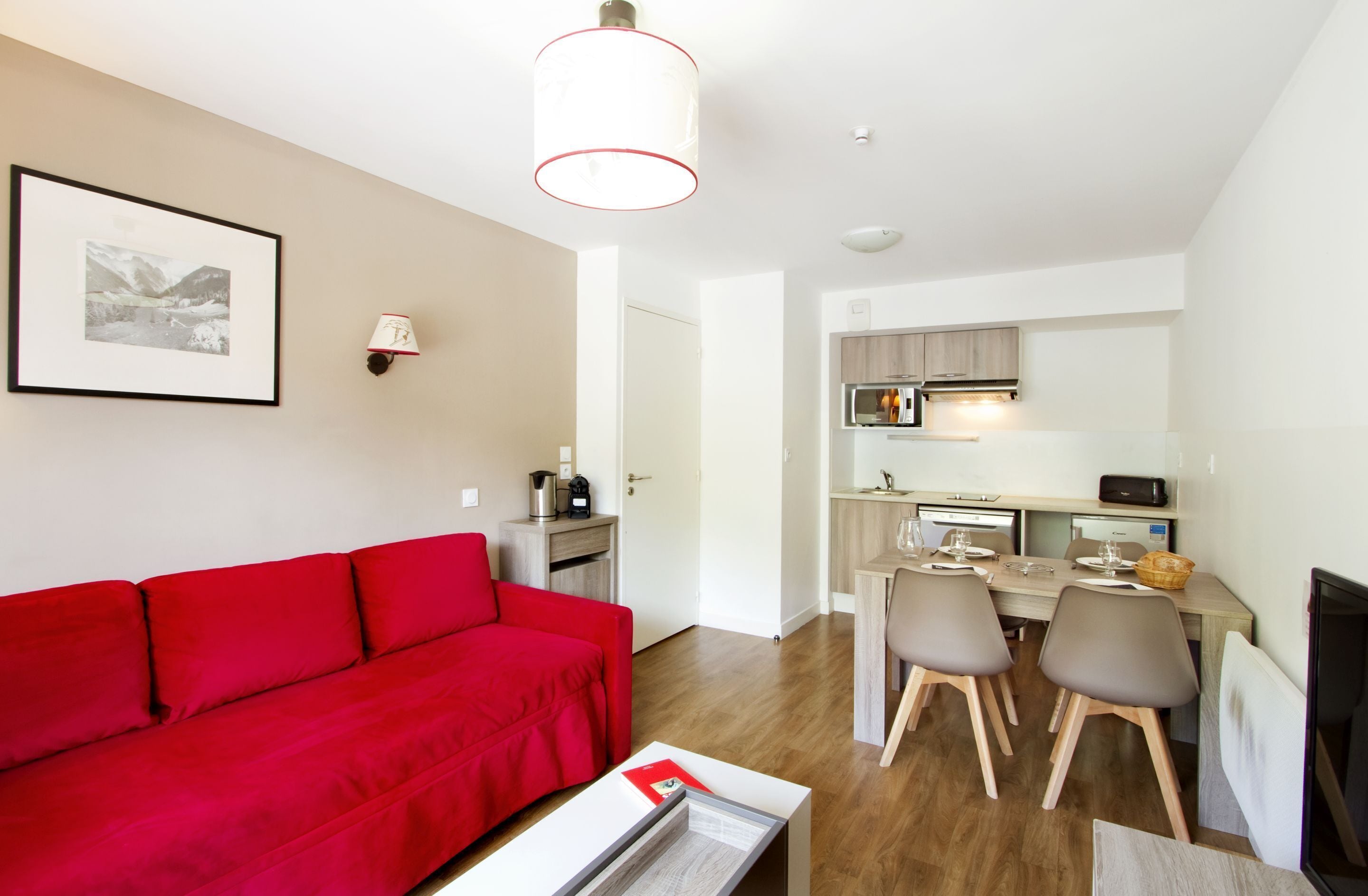 A modern apartment with a red sofa, a dining area with a table and chairs, a kitchen with appliances, and a framed picture on the wall.